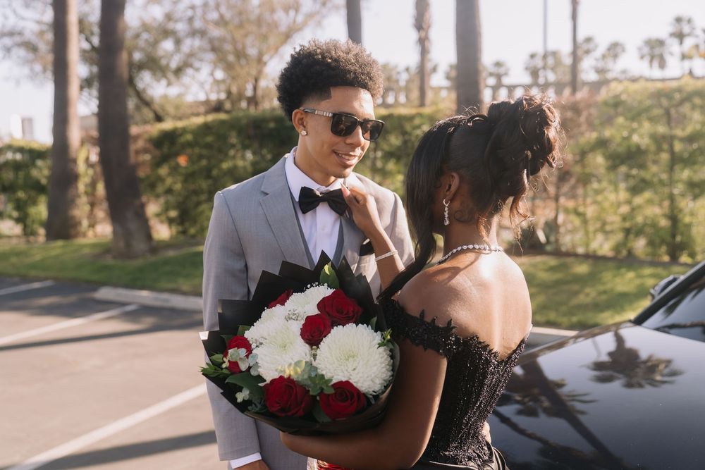 Young couple dressed for prom, boy in gray suit holding red and white bouquet, girl in black off-shoulder gown outdoors.