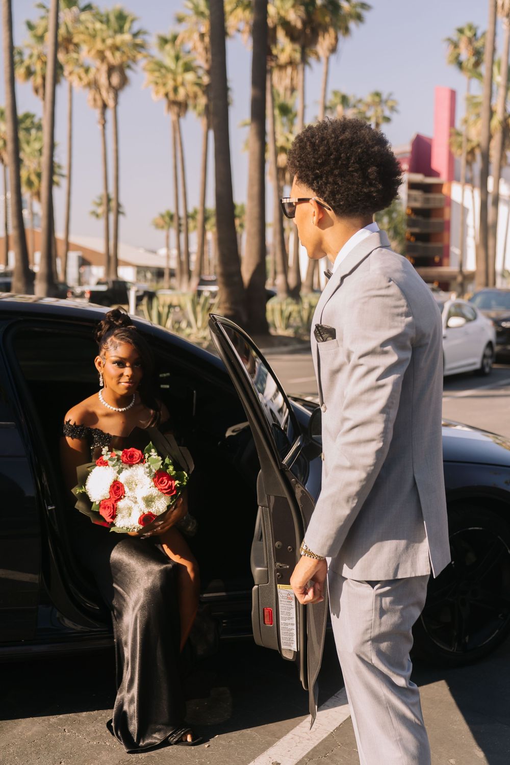 Elegant couple at prom, man in gray suit opening car door for woman in black gown holding bouquet.