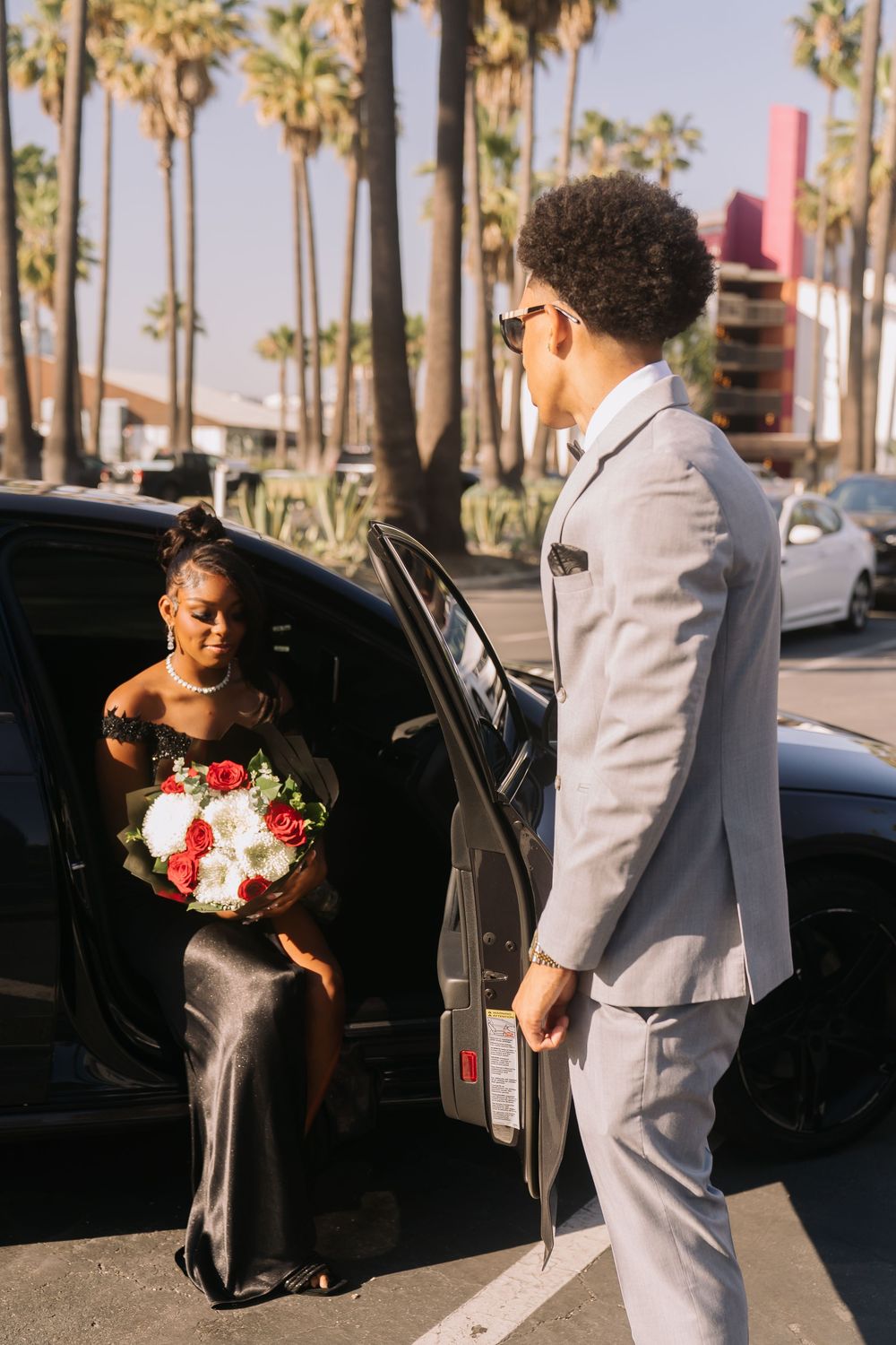 Young man in gray suit opening car door for woman in black dress holding red and white bouquet.