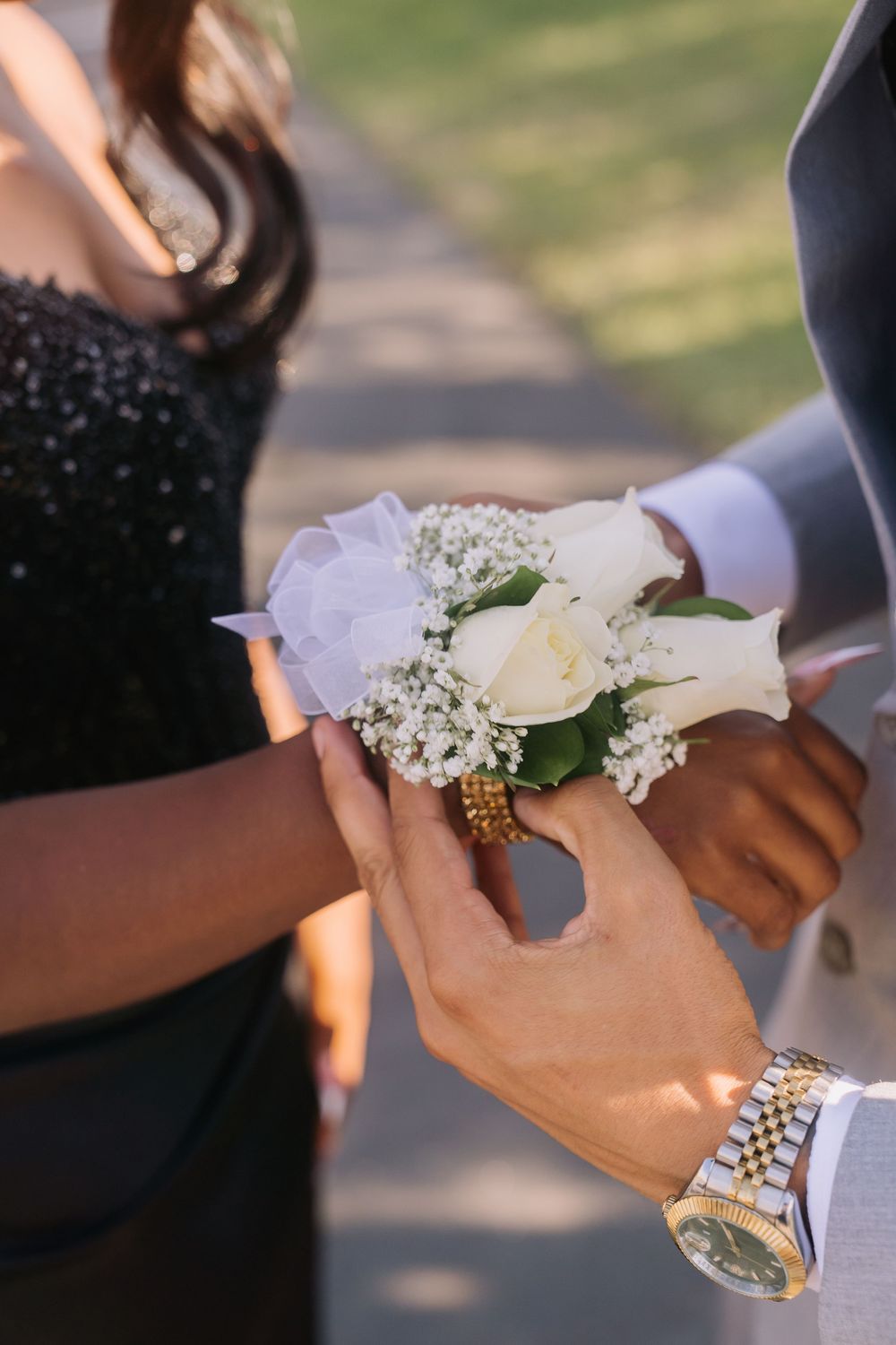 Young man placing a white rose wrist corsage on his prom date's wrist, wearing a gold Rolex watch.
