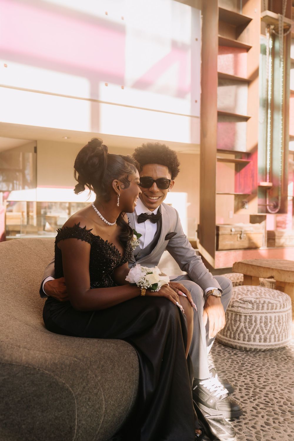 Elegant prom couple sitting together indoors, young man in gray suit with sunglasses and girl in black lace gown smiling.