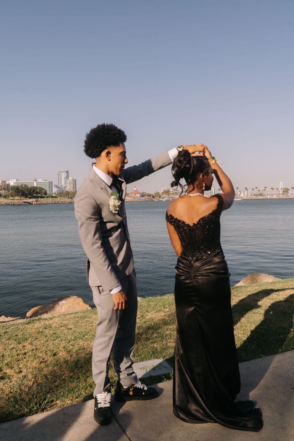 Young prom couple in formal wear standing by the waterfront, man in gray suit adjusting woman's updo hairstyle.