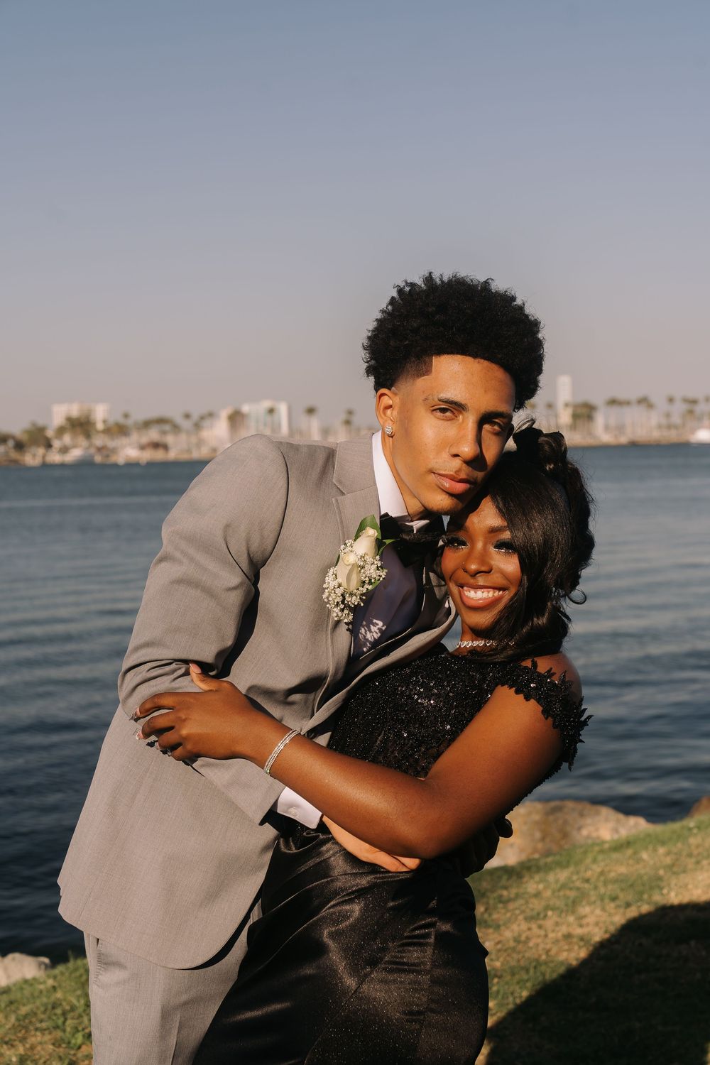 A young couple in prom attire poses by the water, he in a gray suit and she in a black sequined dress.