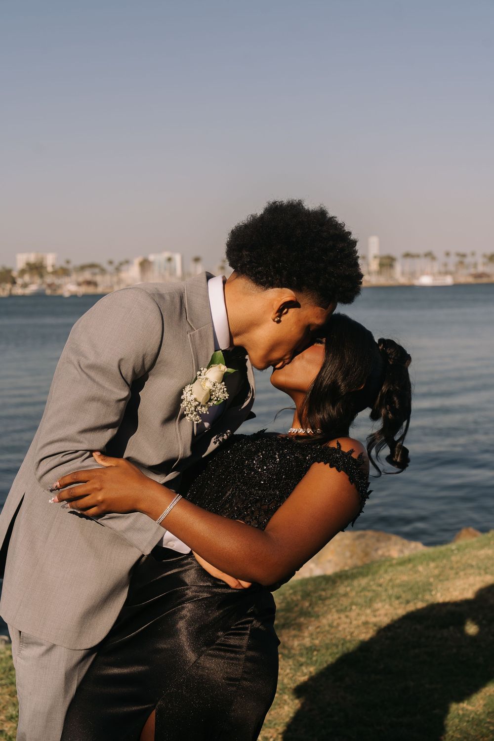Couple sharing a romantic moment outdoors near the water, man in gray suit leaning in to kiss woman in black dress.
