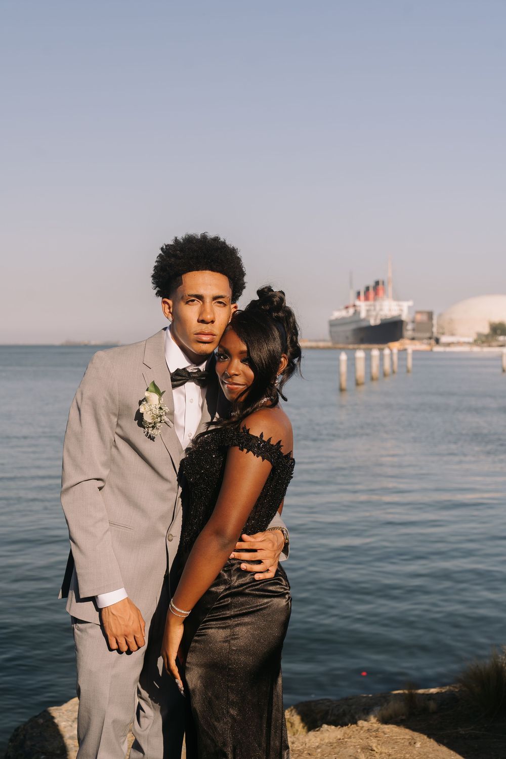 Young couple in formal wear posing by the waterfront, man in gray suit with boutonniere and woman in black lace dress.
