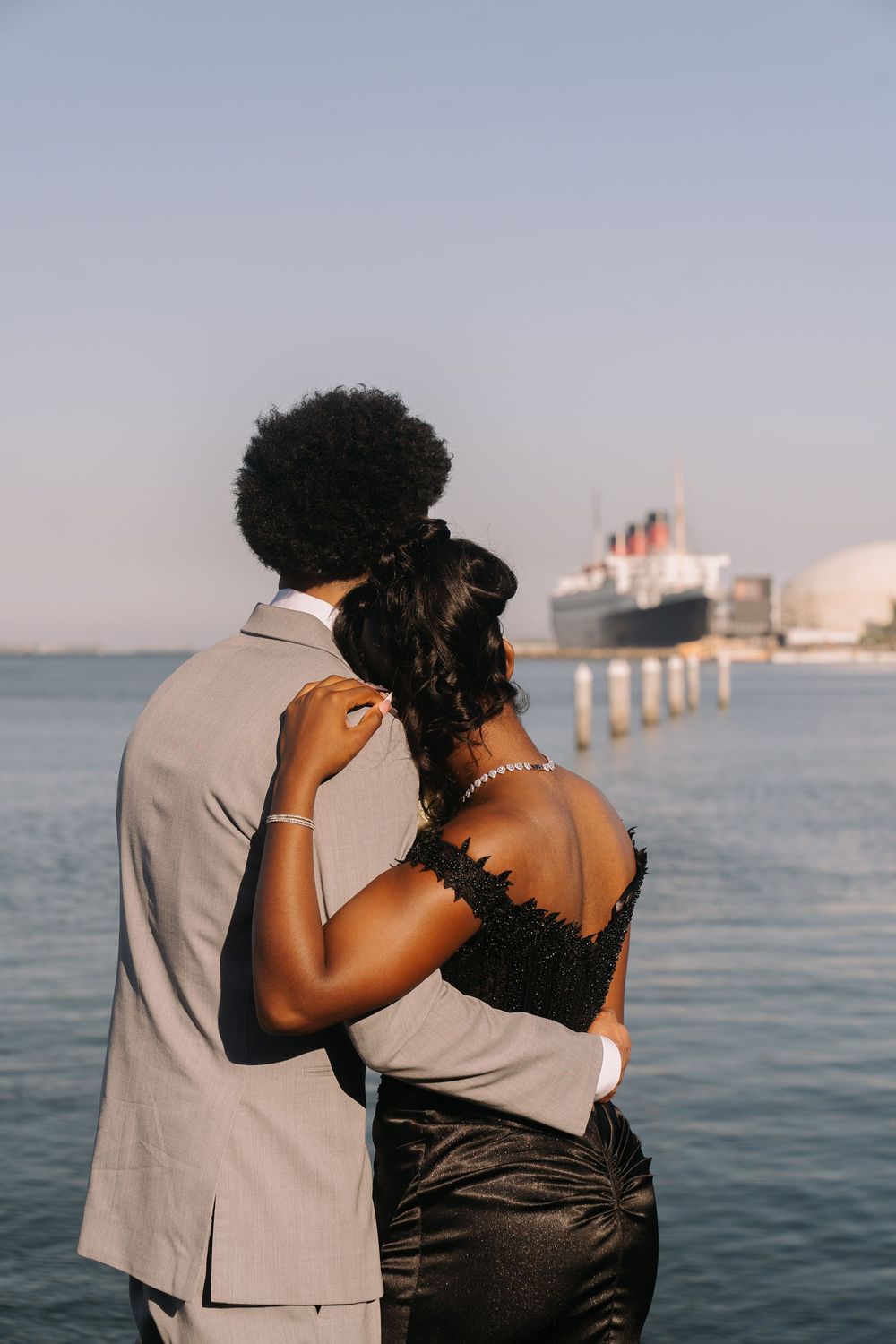 Prom couple in formal attire embracing while gazing at a ship on the water during golden hour.