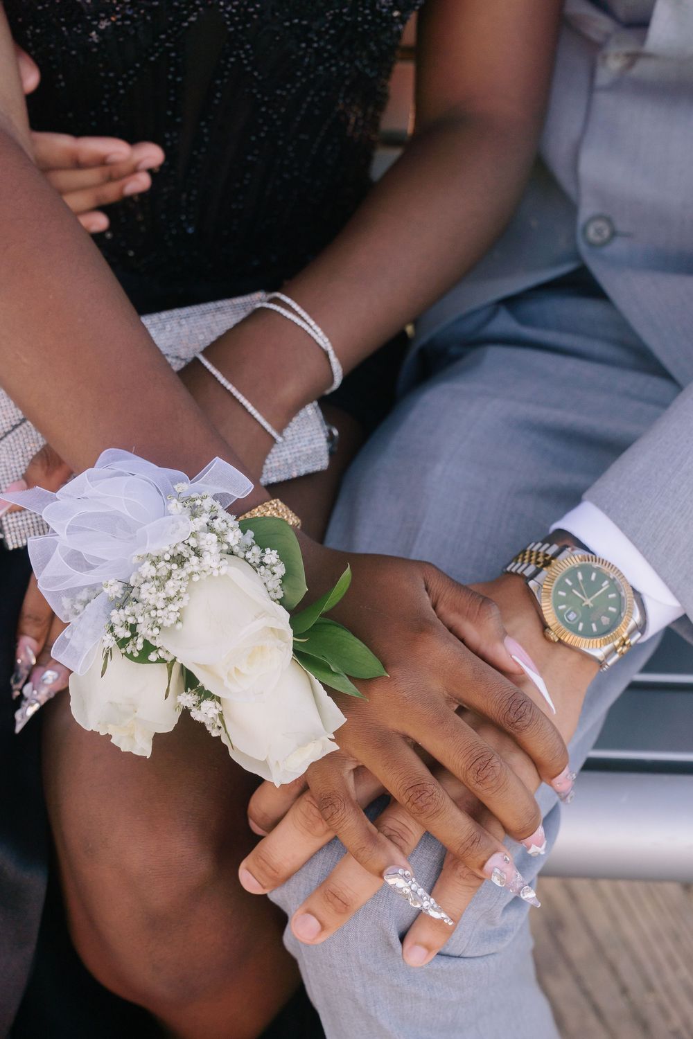 Couple holding hands at prom, woman wearing white rose wrist corsage, man wearing gold watch, both dressed formally.