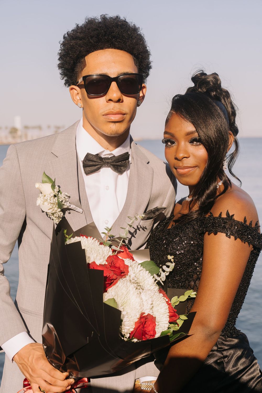 A stylish prom couple poses outdoors, he in a gray tuxedo with sunglasses holding a red and white bouquet.