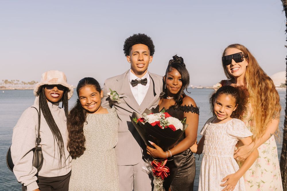 Group of seven people posing together outdoors near water at a formal event, with one person holding a flower bouquet.