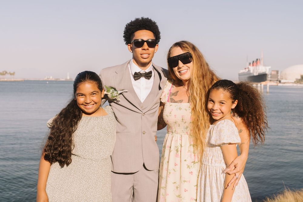 Family of four posing outdoors near waterfront, two adults wearing sunglasses with two young girls in formal attire.