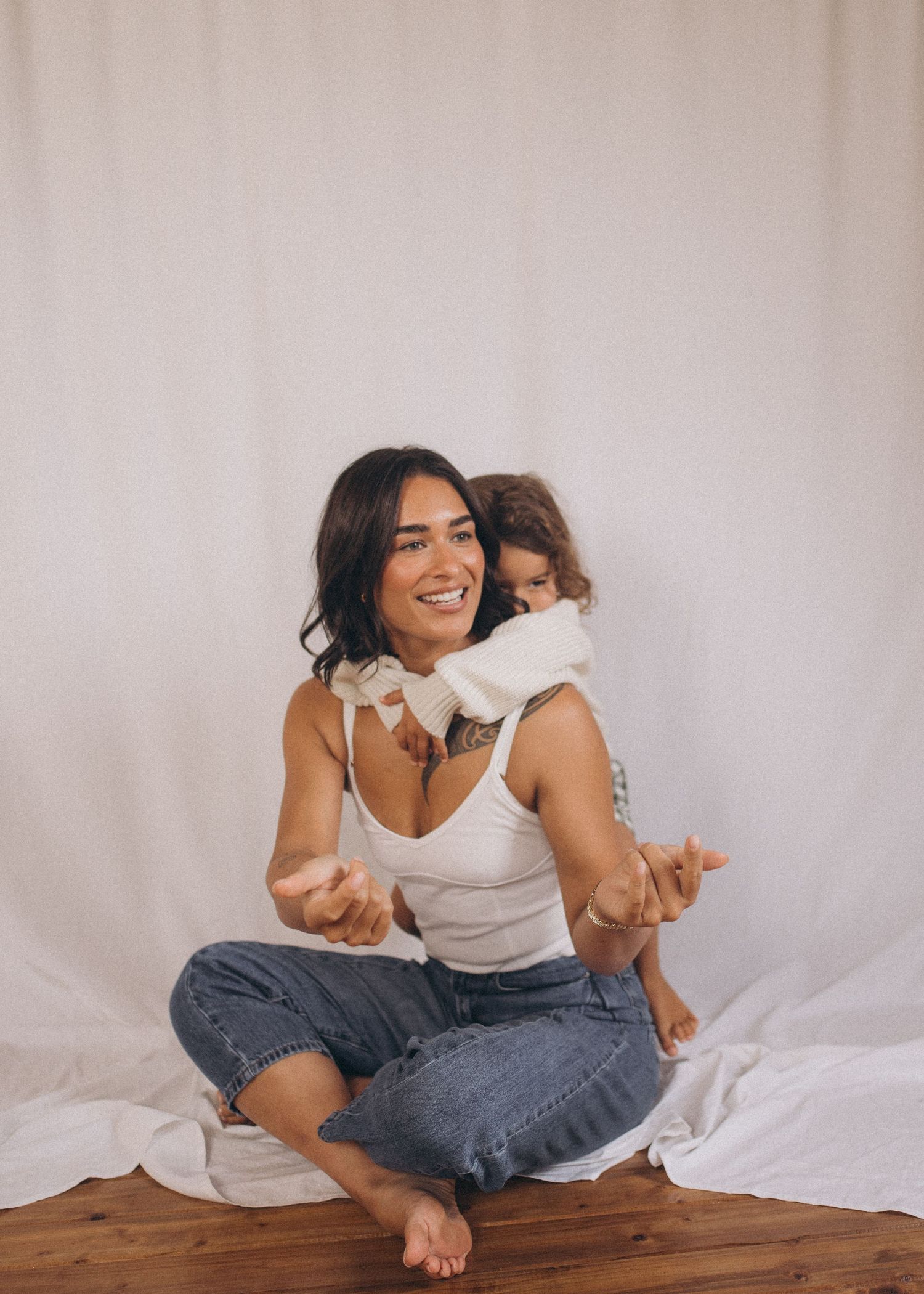 A smiling woman in white tank top and jeans sits cross-legged on wooden floor while a toddler hugs her from behind in a portrait studio.