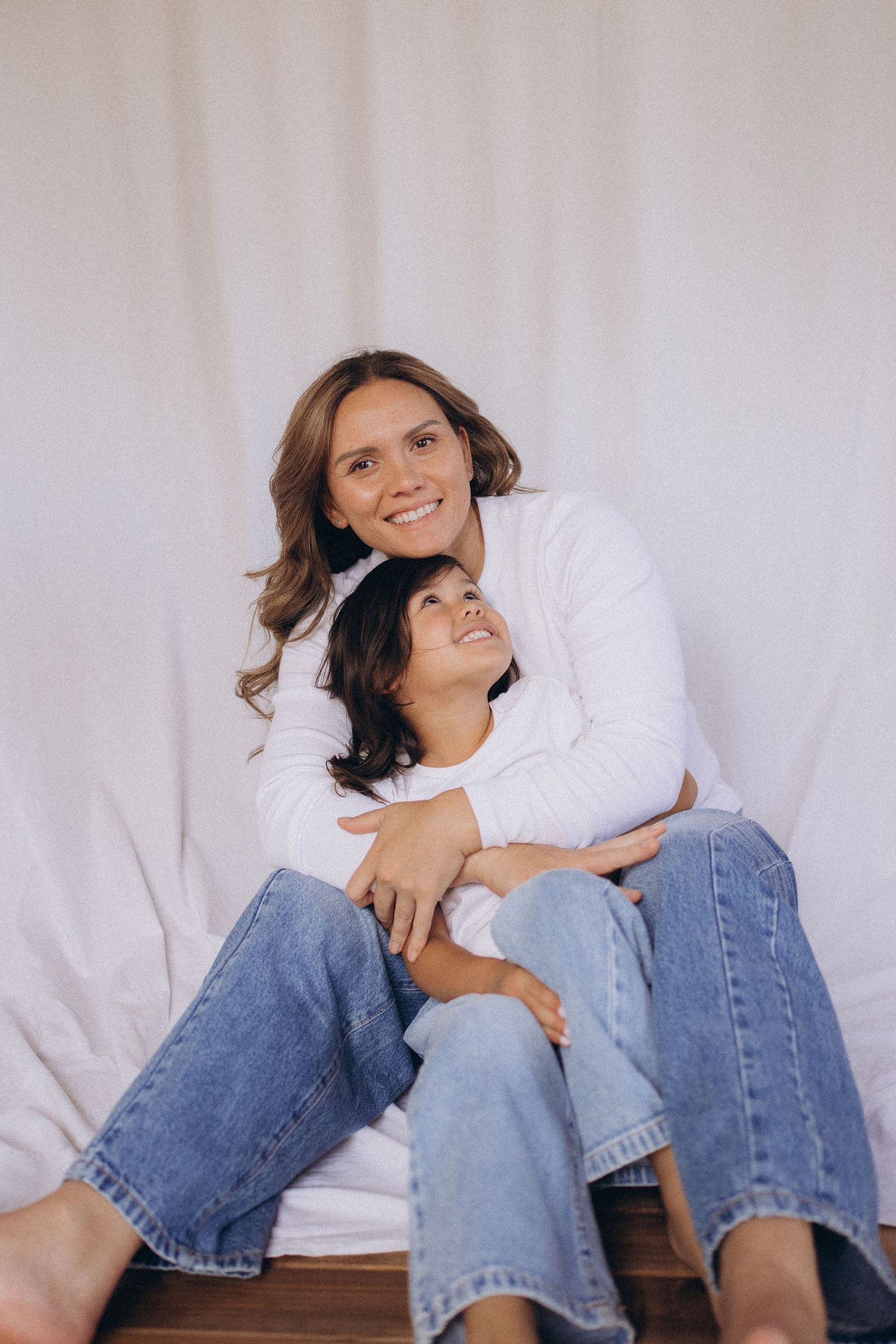 A happy mother and child sit together on a wooden floor, embracing warmly against a white fabric background in New Zealand portrait studio.