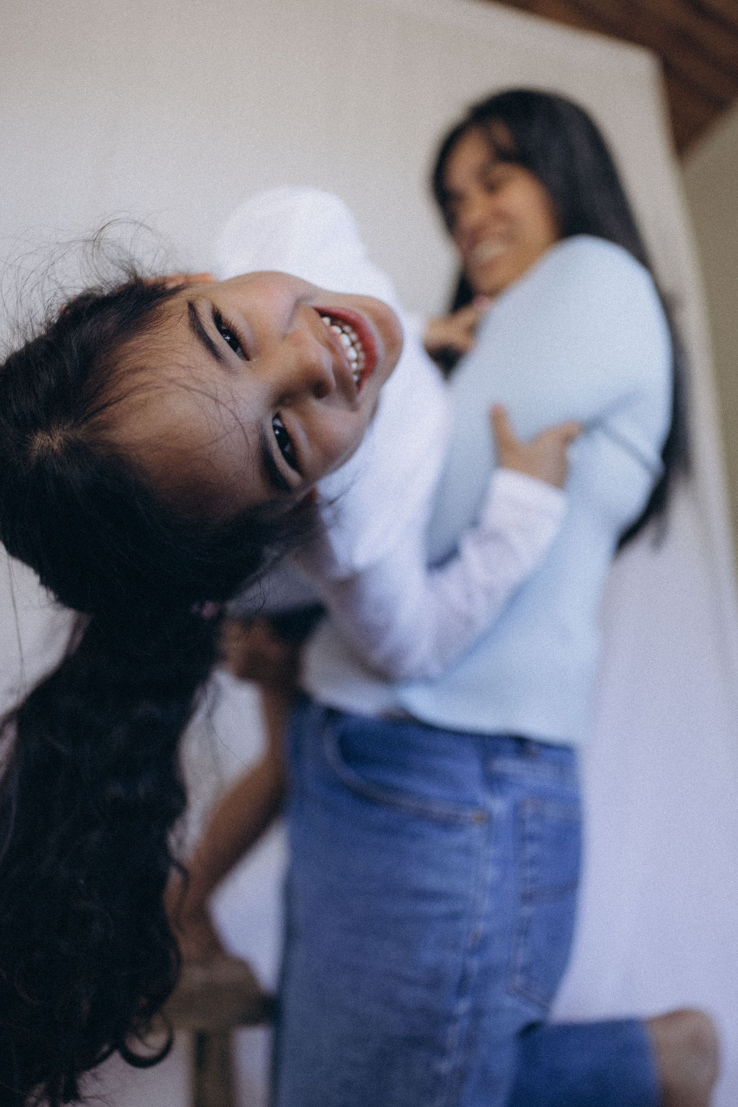 Joyful young girl with long dark hair laughing while a woman holds a child in the background in New Zealand..