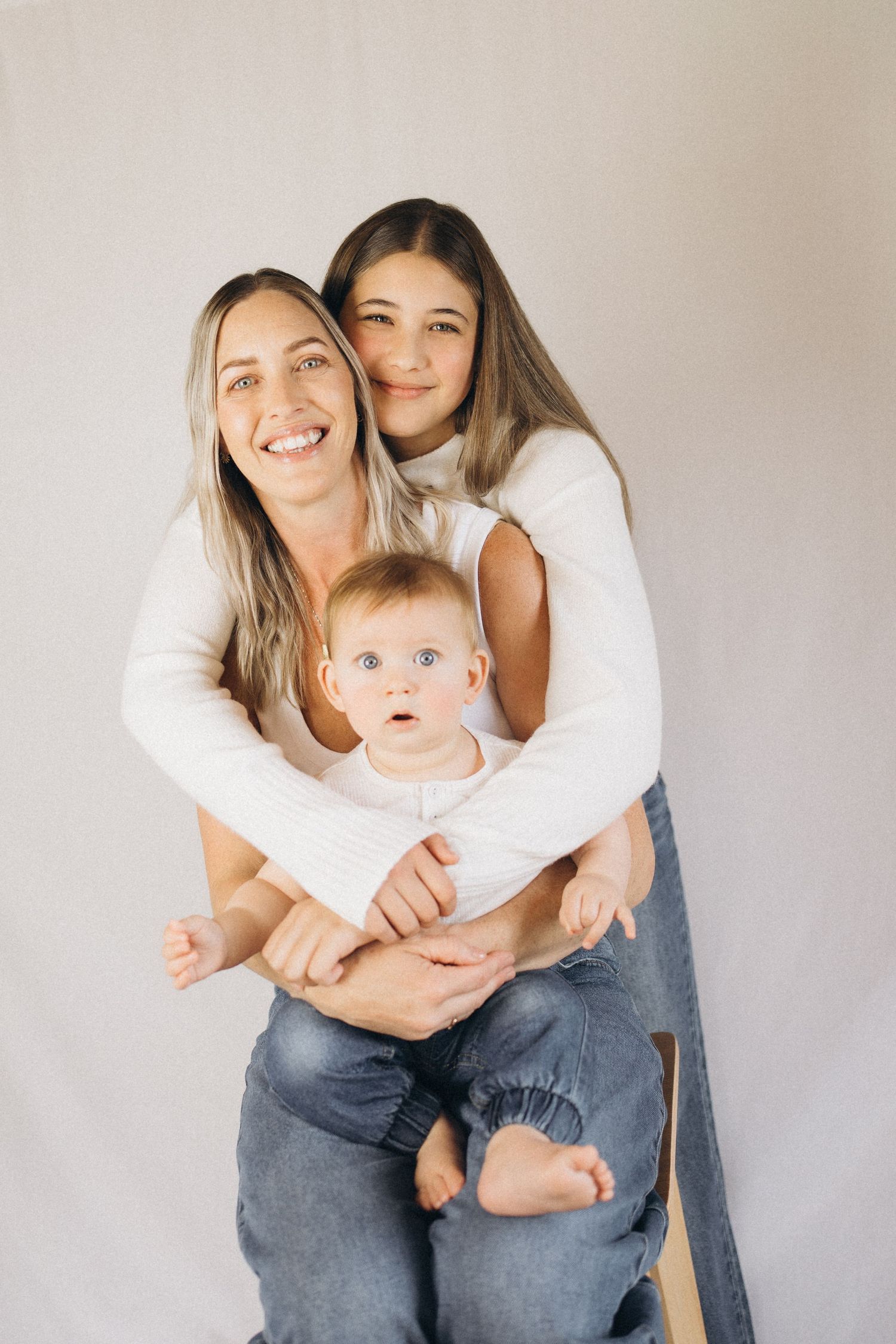 Smiling mother with blonde hair holds a baby while her teenage daughter embraces them from behind in New Zealand..