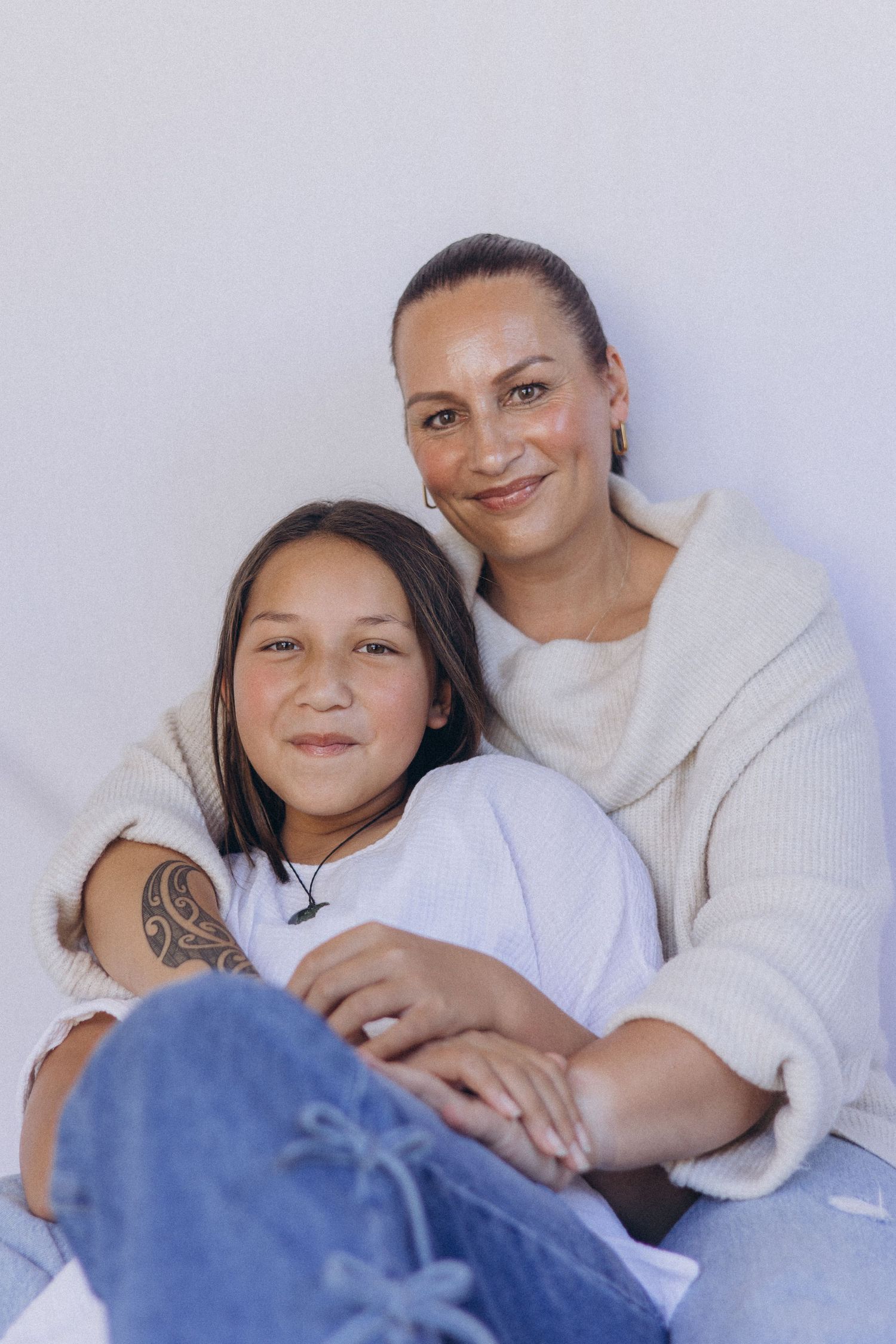 A smiling mother and her young daughter with a tattoo sit together against a white wall in New Zealand.