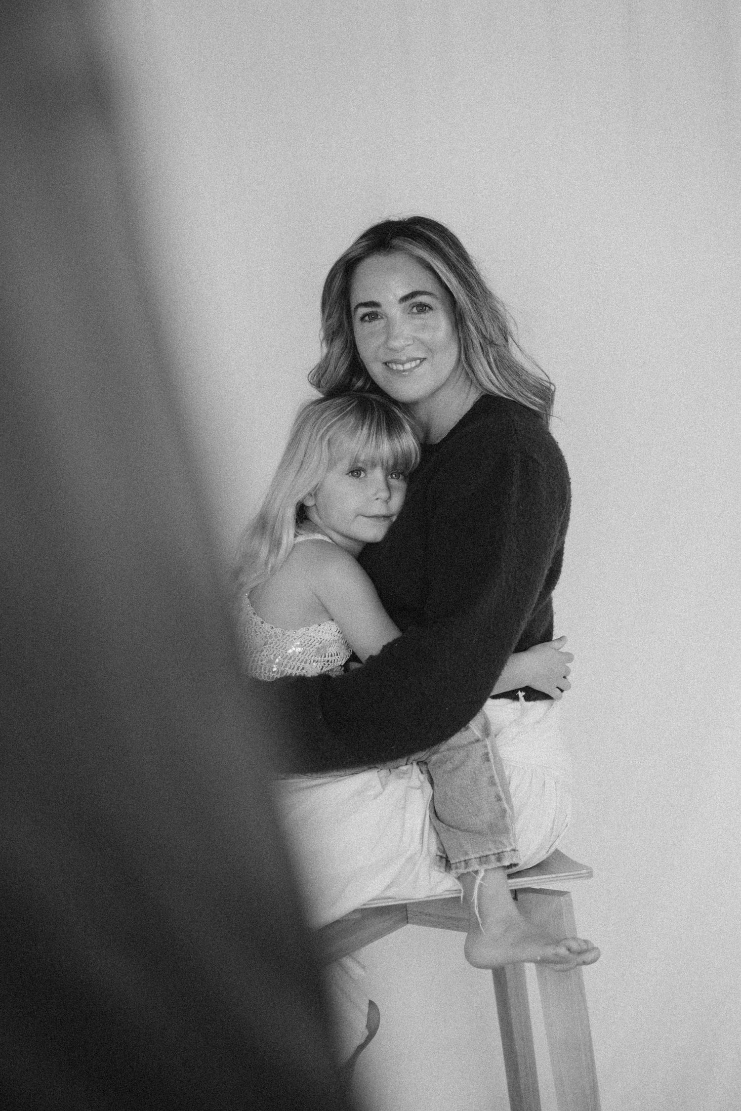 Black and white portrait of a mother embracing her blonde daughter on a stool against a light wall in New Zealand..