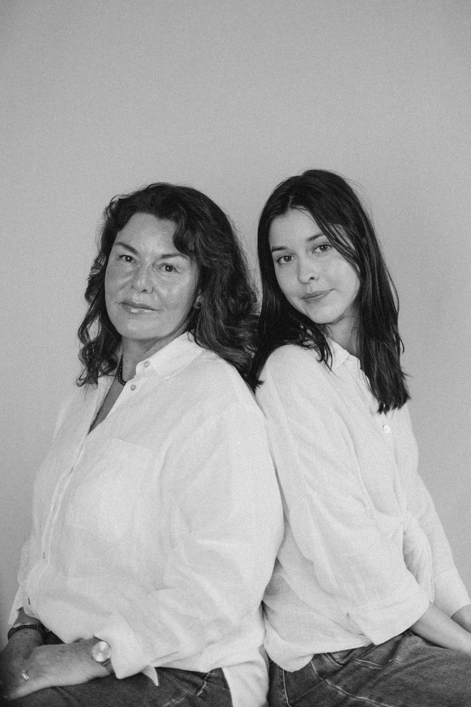 Black and white portrait of two women in white linen shirts sitting back to back against a neutral wall in New Zealand..