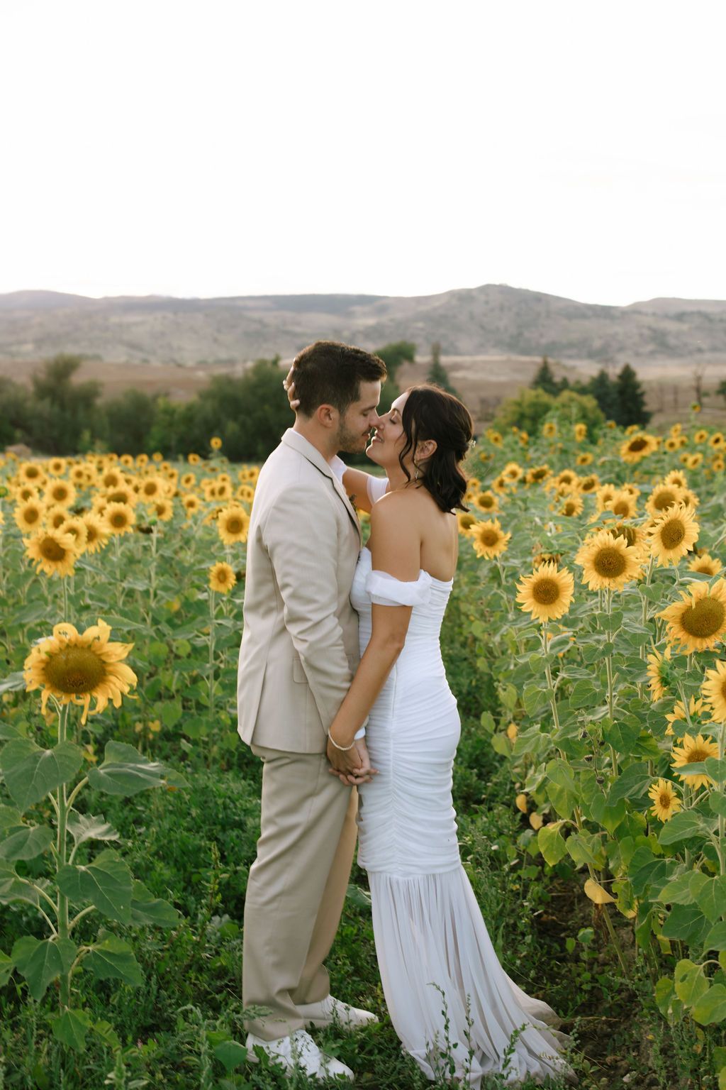 Bride and groom embracing in a sunflower field, wearing white gown and beige suit, with mountains in the background.