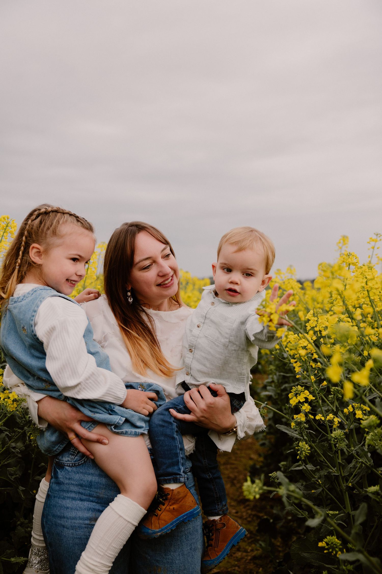 Smiling mother holding two young children in a blooming yellow rapeseed field outdoors.