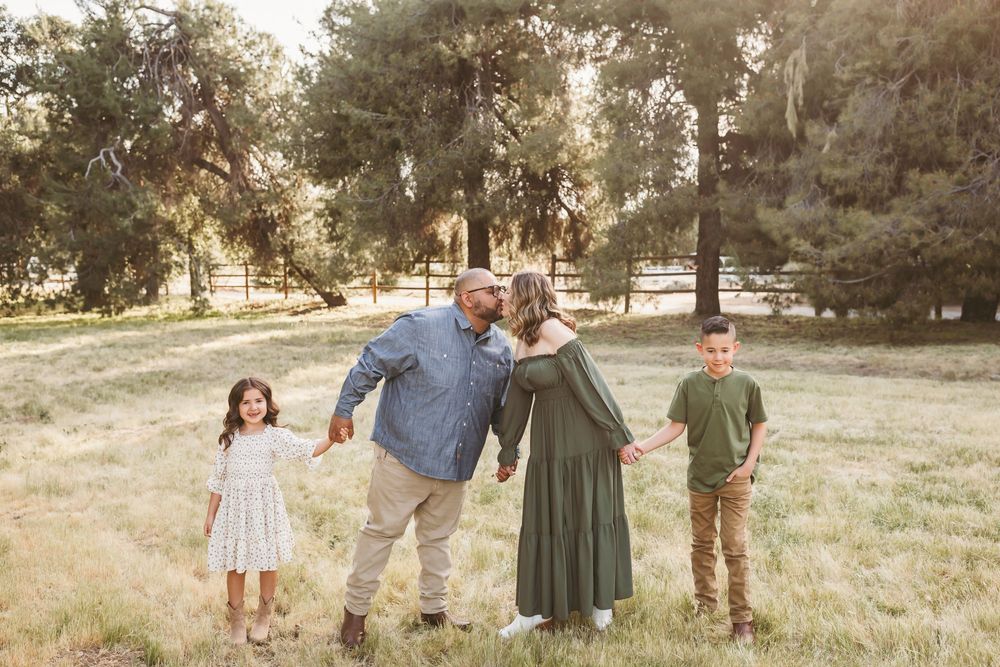 Family of four poses in a sunlit meadow with tall trees, parents kissing while holding hands with their two young children in the inland emp