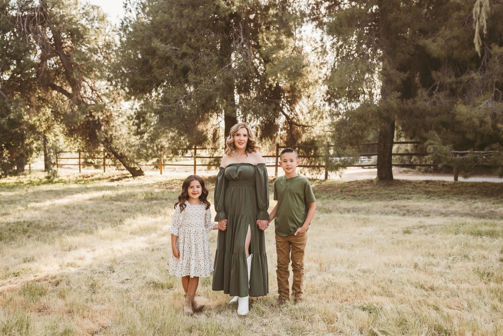 Pregnant woman in green dress poses outdoors with two children in a sunny park in yucaipa ca