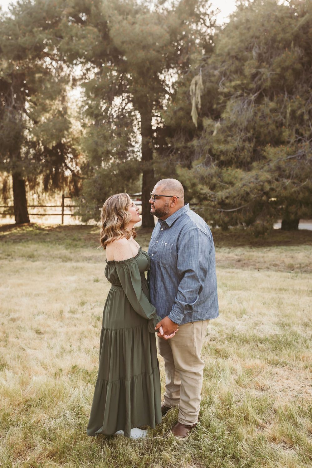 Couple holding hands and gazing at each other in a sunlit field, woman in olive dress, man in denim shirt.