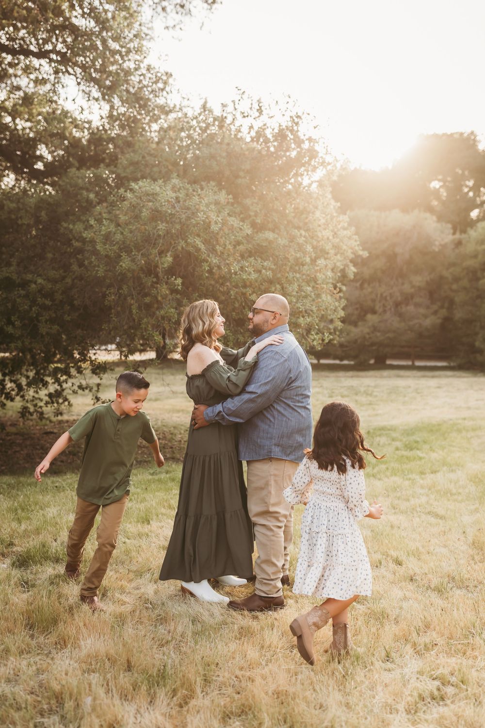 Family of four sharing a warm embrace in a sunlit meadow, with children running toward their parents at golden hour.