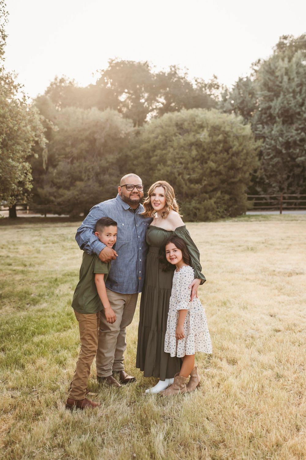 Family of four posing together in golden hour field, parents with two children in coordinating green outfits.