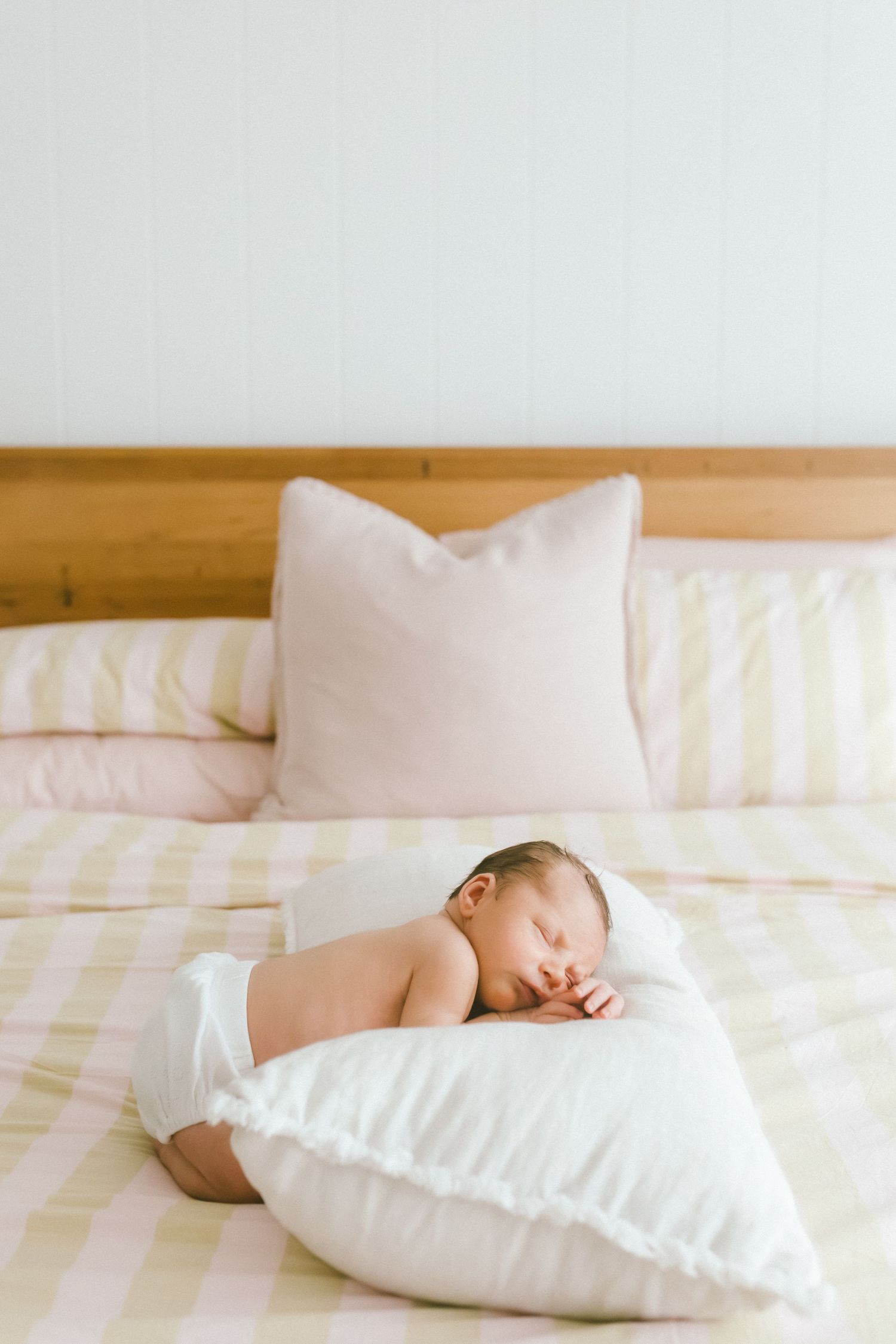 Newborn baby sleeping peacefully on a white pillow on a pink striped bed with wooden headboard.