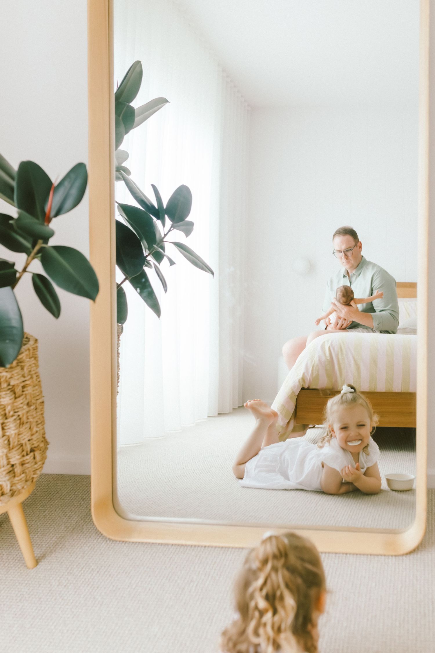 Family enjoying a bright bedroom, with a father holding a baby on the bed and a toddler smiling in a large mirror reflection.