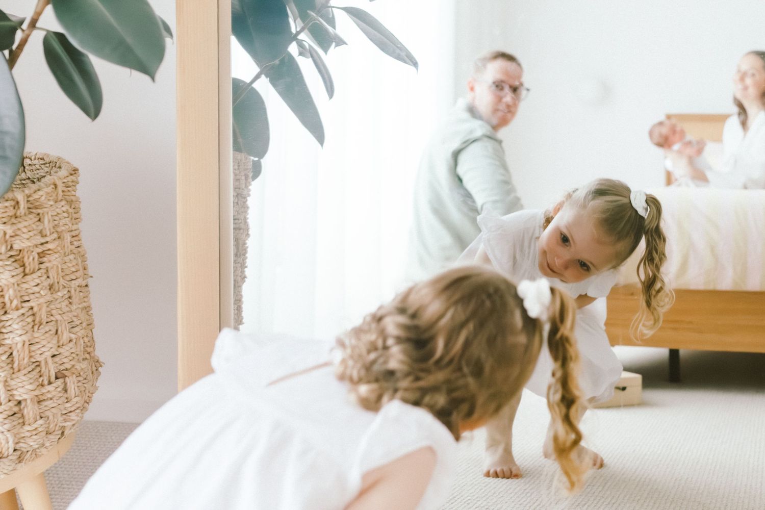 Yong girl looking in the mirror & playing on a bright bedroom floor while parents relax on a bed in a modern home.