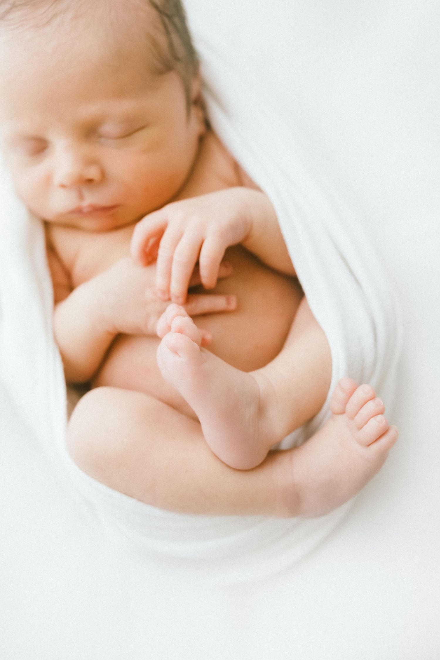 Peaceful sleeping newborn baby wrapped in white swaddle blanket with tiny hands and feet curled up.