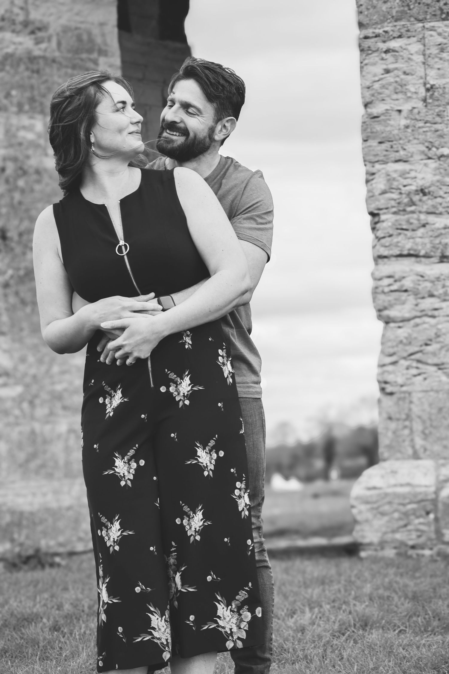Black and white photo of a smiling couple embracing outdoors near stone ruins, woman in floral dress, man in t-shirt.