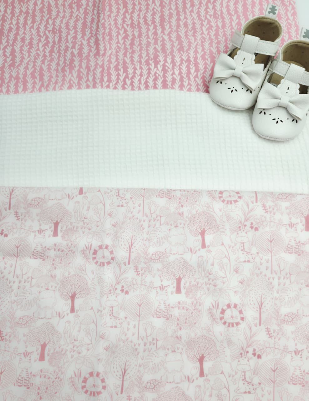 Pink and white baby blanket with woodland print, displayed flat with white bow Mary Jane baby shoes on top.