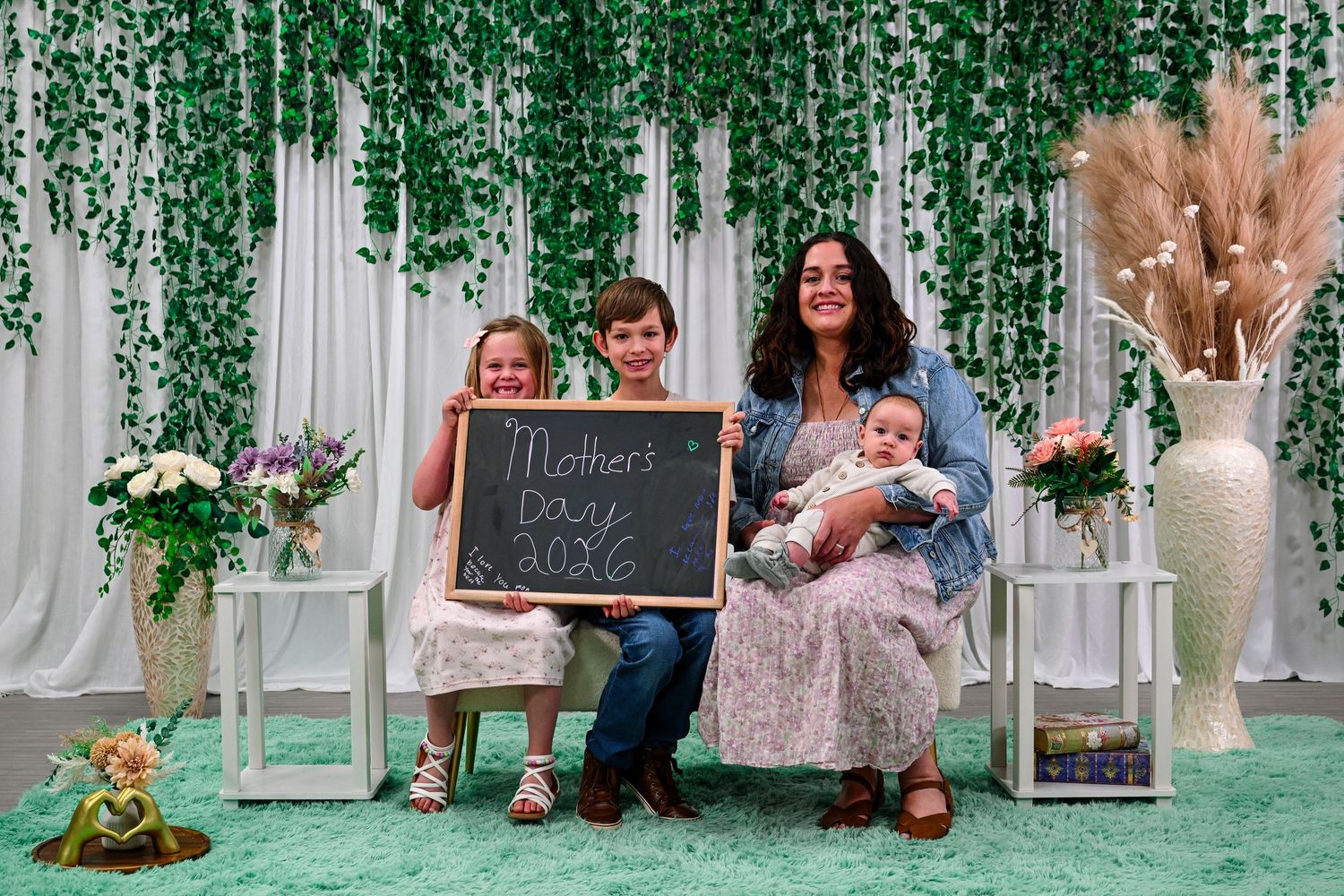 Family of five posing for a Mother's Day 2026 photo with a chalkboard sign, greenery backdrop, and floral decor.