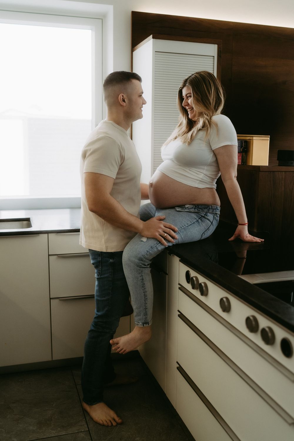 Pregnant woman sitting on kitchen counter smiling at partner in modern kitchen, both wearing white shirts and jeans.