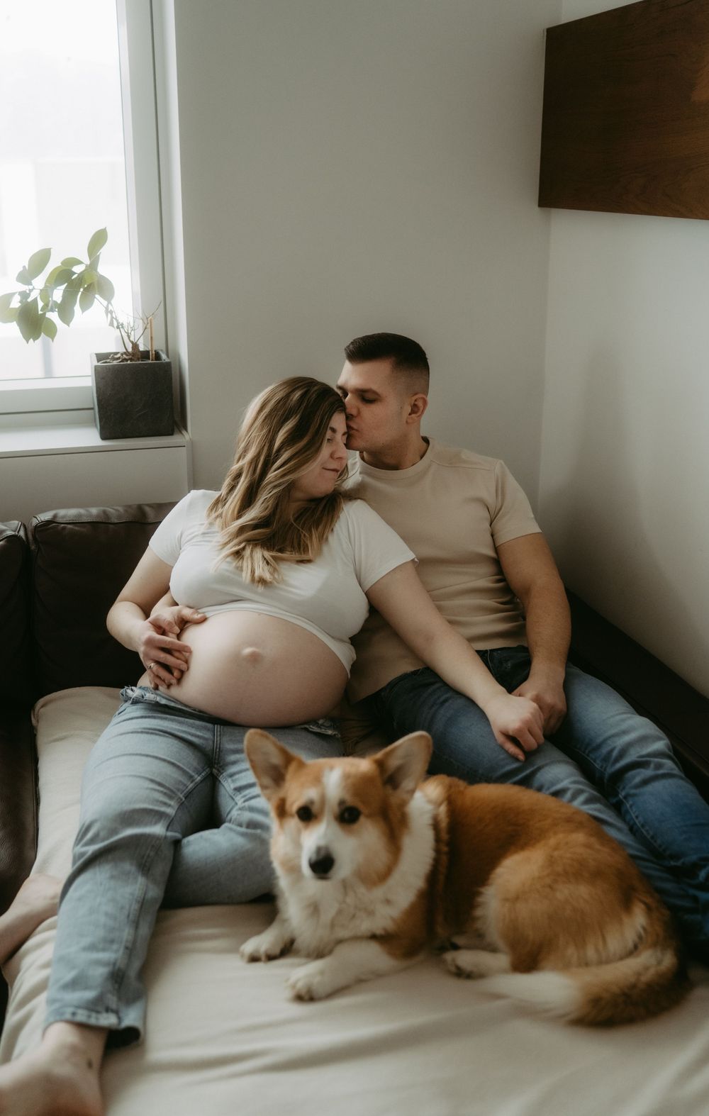 Pregnant couple sitting on couch with Corgi dog, sharing tender kiss in cozy home setting.