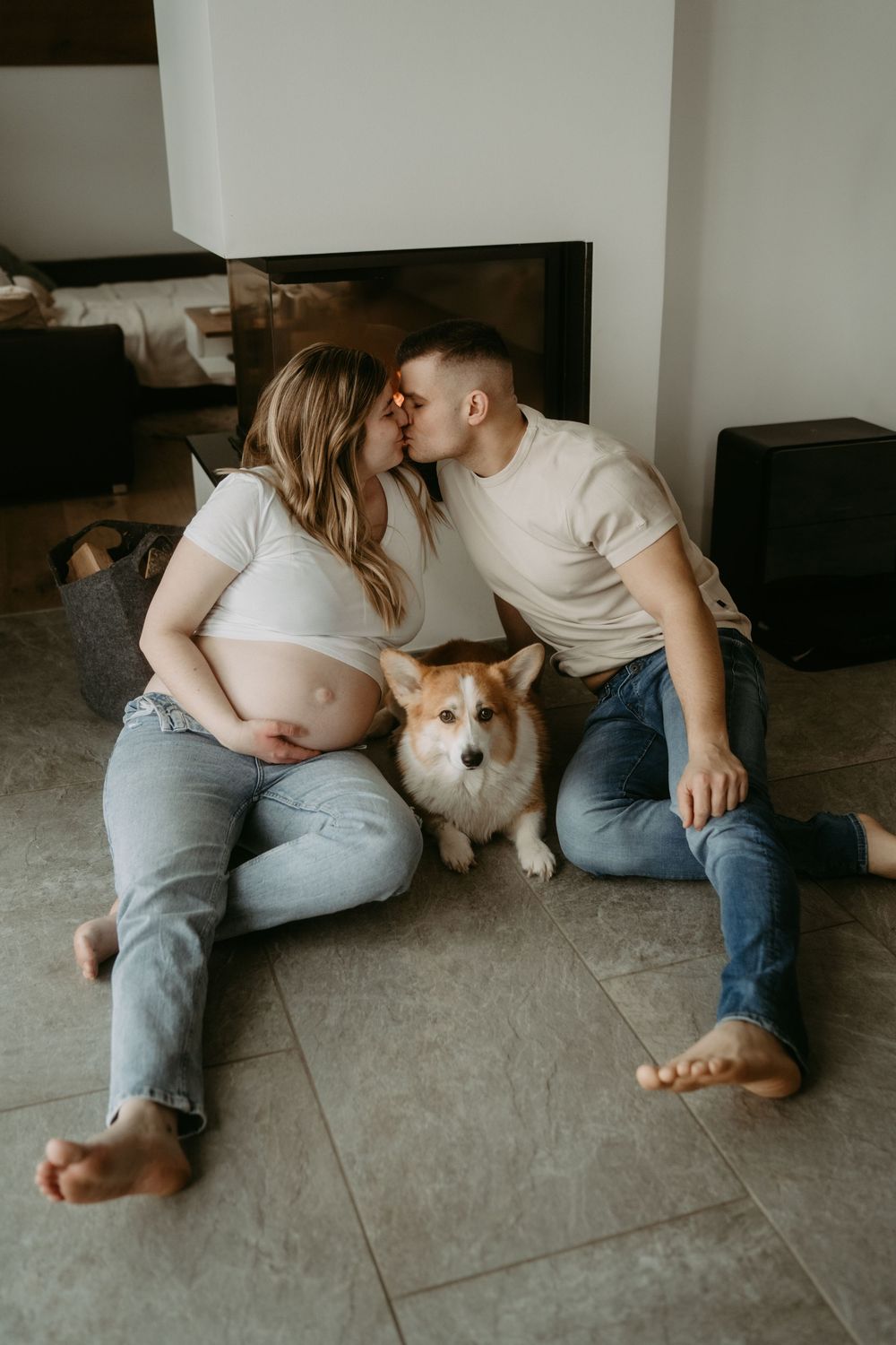 Pregnant couple kissing on floor with corgi dog sitting between them near fireplace.