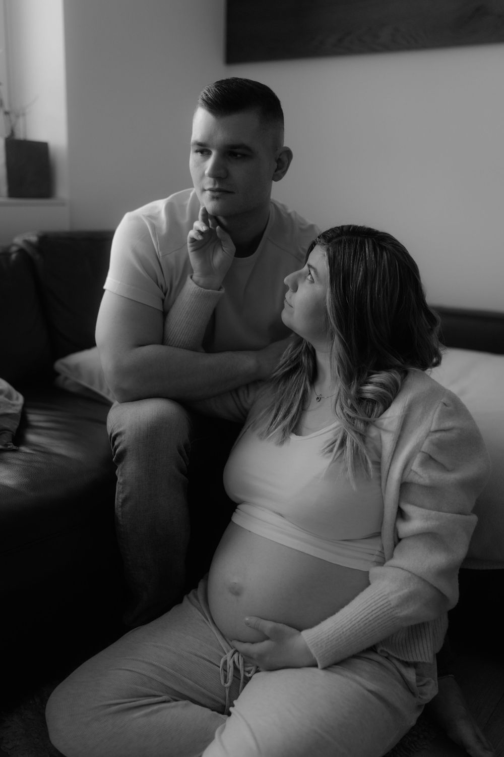 Black and white photo of expectant couple relaxing on sofa, woman cradling pregnant belly while partner sits behind.