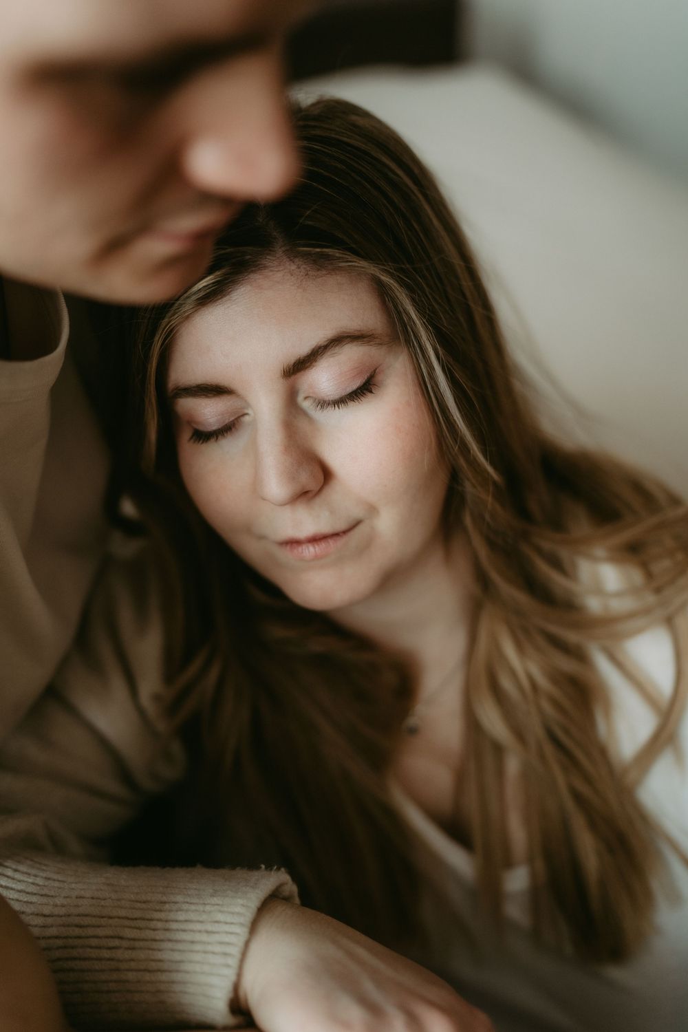 Close-up of pregnant woman with long hair, eyes closed, leaning against partner in soft light.