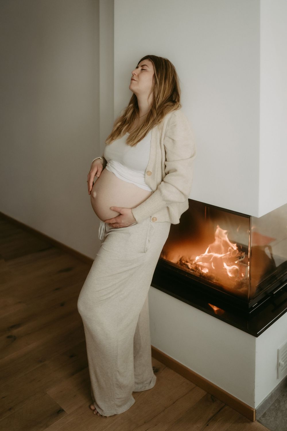 Pregnant woman in beige outfit standing by lit fireplace cradling baby bump.