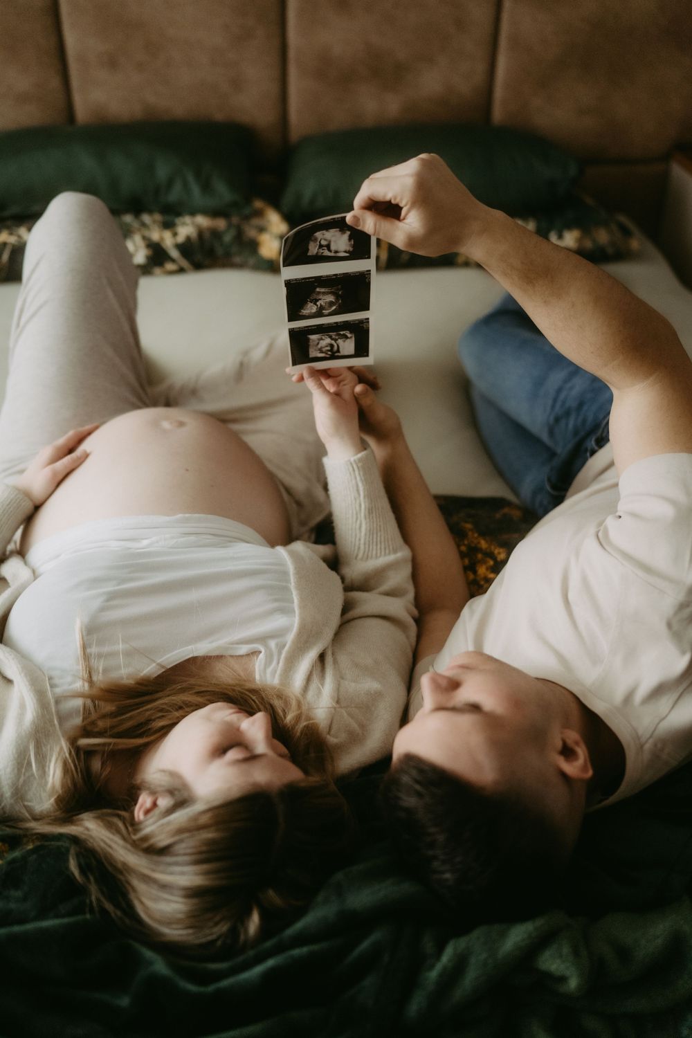 Pregnant couple lying on bed looking at ultrasound photos together in cozy bedroom setting.