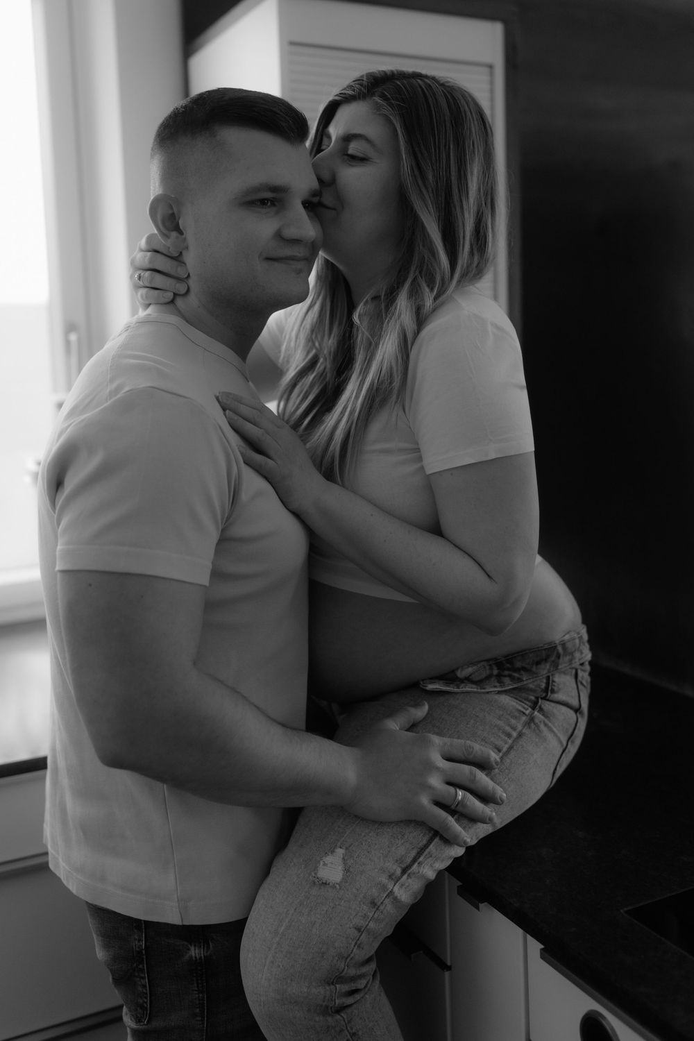 Black and white photo of a couple embracing in a kitchen, woman kissing mans cheek tenderly.