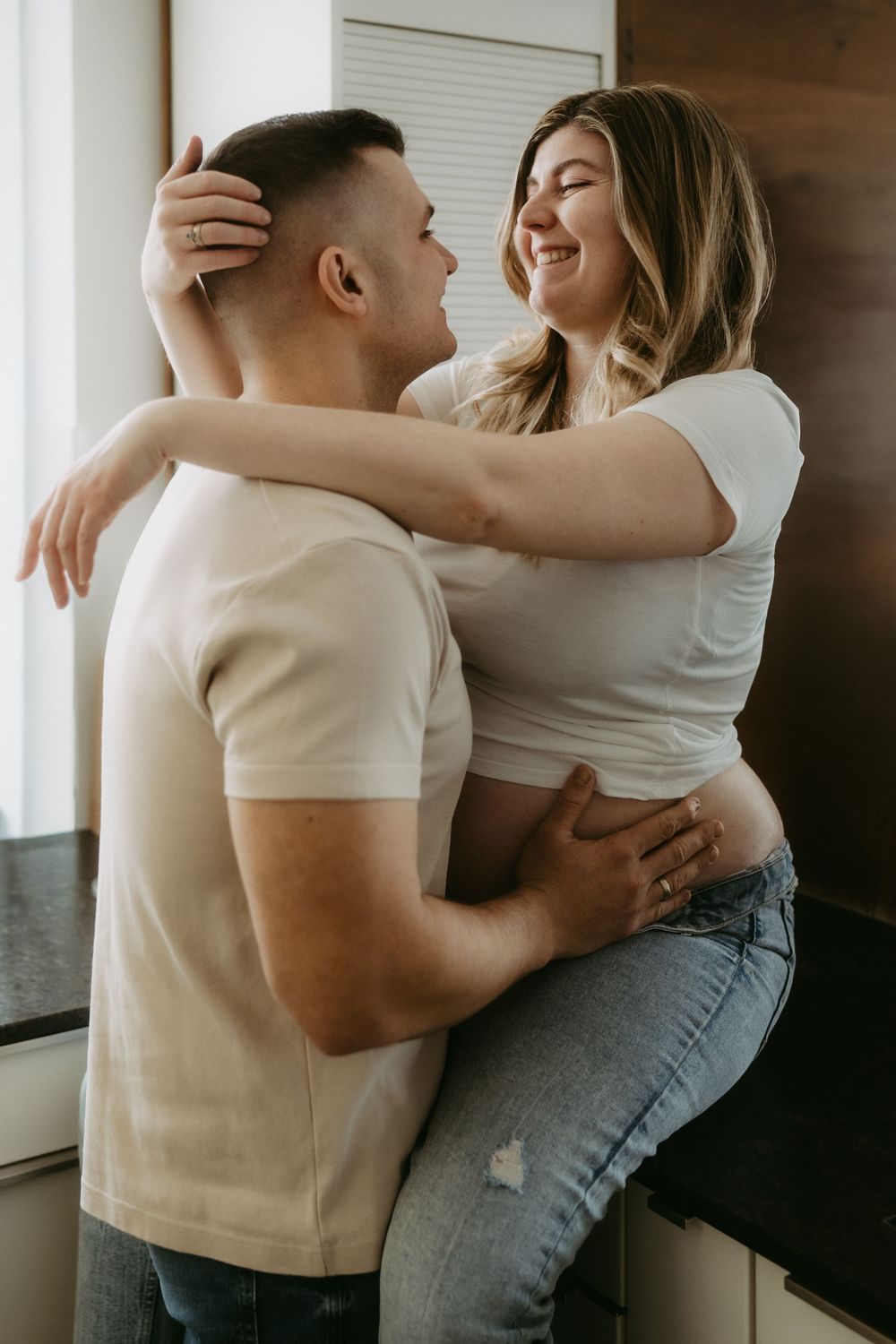 Happy pregnant couple embracing and smiling at each other in a bright kitchen setting.