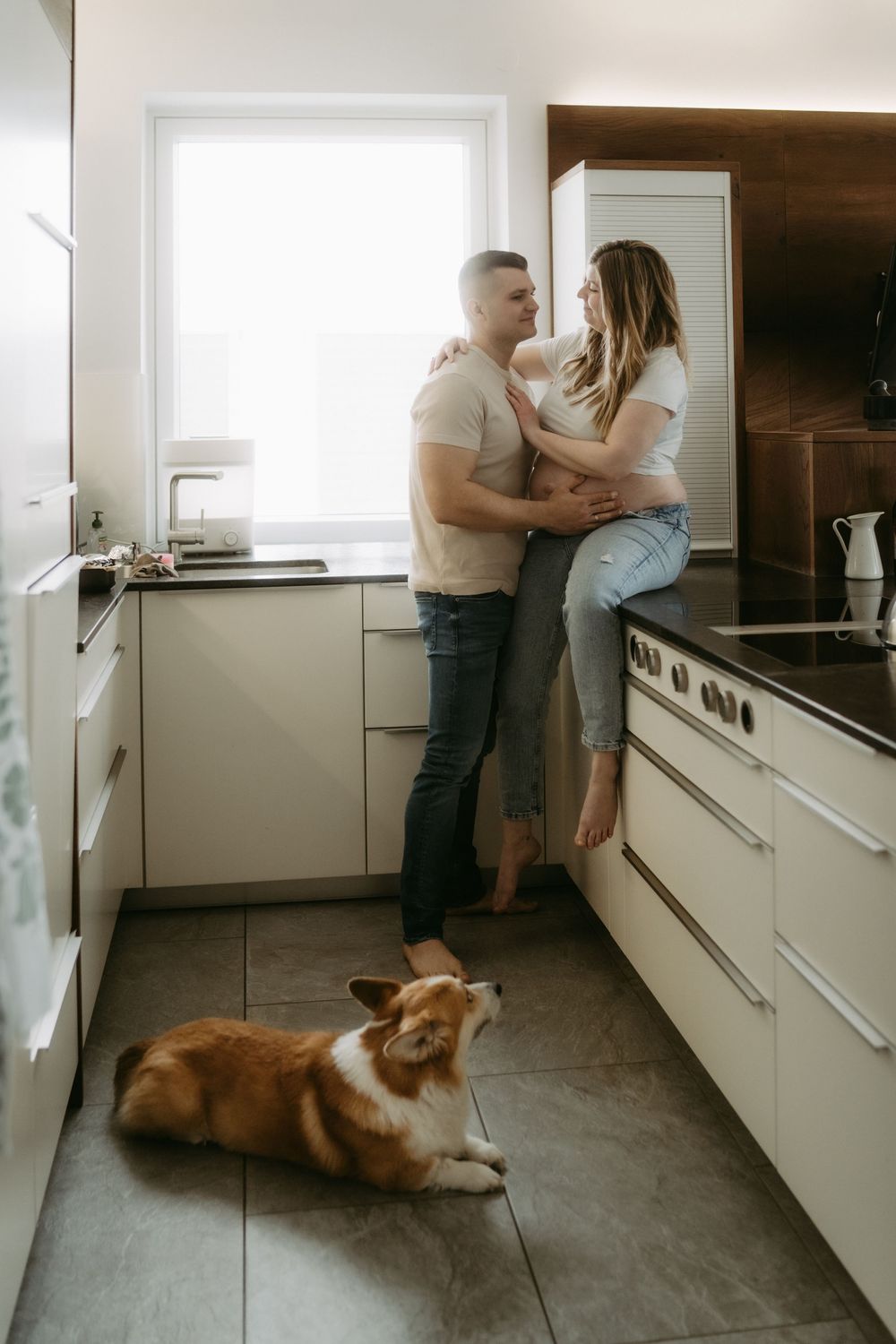 Couple sharing a romantic moment in a modern kitchen while their Corgi dog rests on the floor.