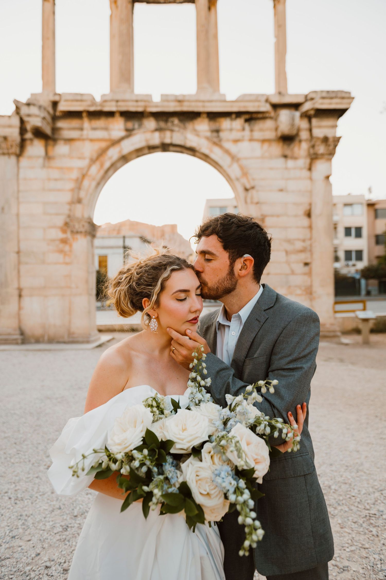 Bride and groom share a romantic kiss in front of Hadrian's Arch in Athens, Greece, with a white floral bouquet.