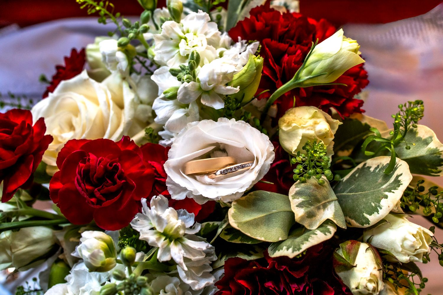 Elegant wedding bouquet featuring red carnations, white roses, and greenery with gold rings nestled among the blooms.