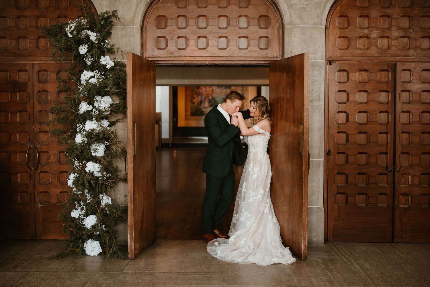 Bride and groom share a romantic kiss in ornate wooden doorway adorned with white floral arch at wedding venue.