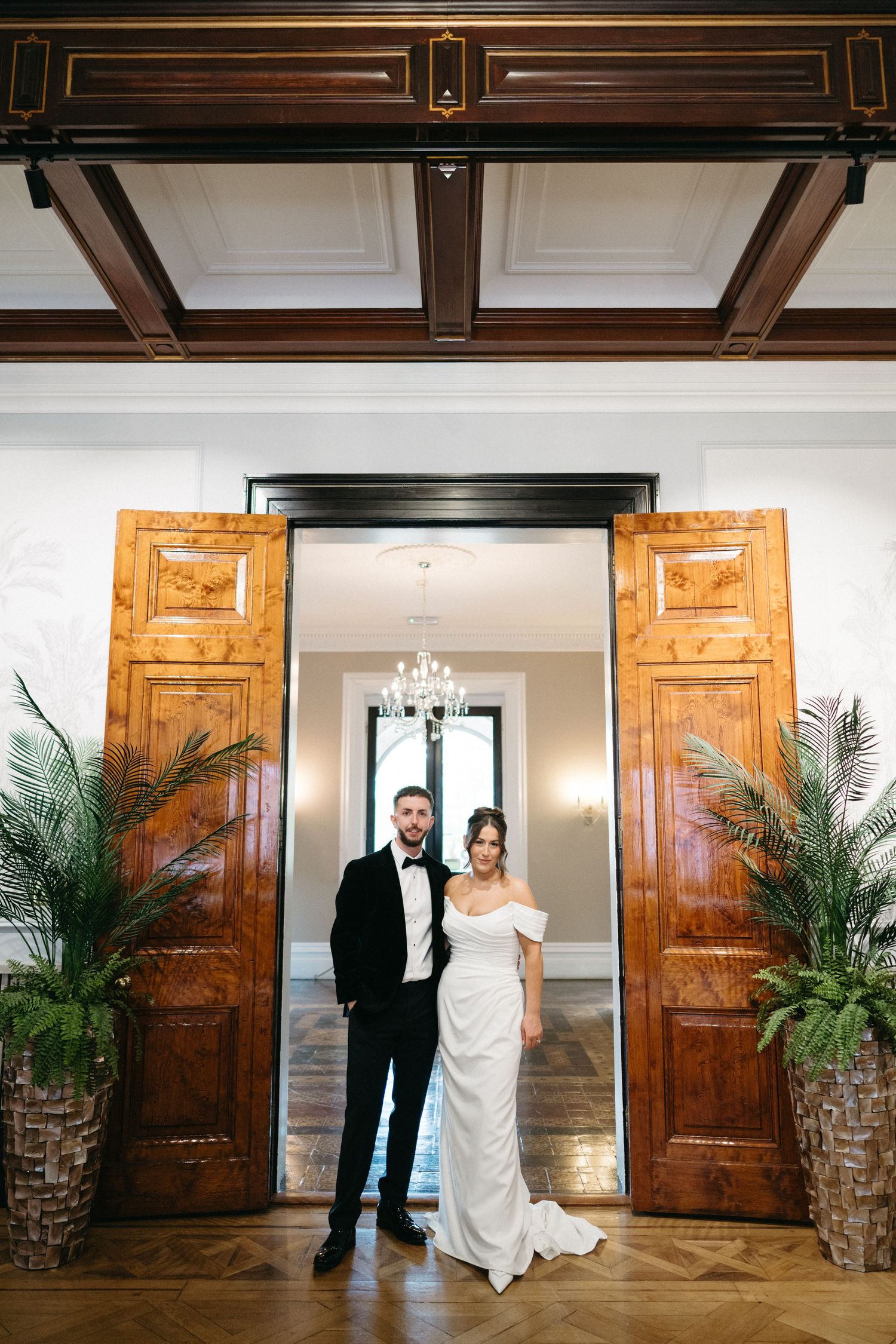Elegant bride and groom posing in front of ornate wooden doors in a grand ballroom wedding venue.