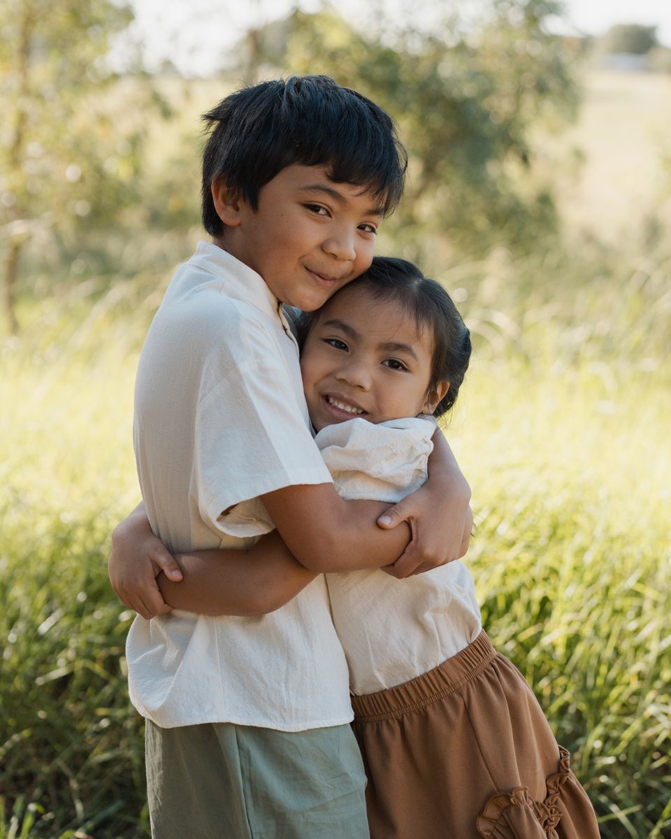 Two young siblings hugging and smiling outdoors in a green field, wearing casual earth-toned clothing during a family photoshoot in sydney